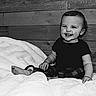 baby, child, smiling, sitting, portrait, black_and_white, blanket, bed, wooden_wall, indoors, happy, grinning, infant, short_hair, black_shirt, barefoot, cozy, rustic, photography, single_person