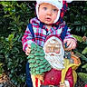 child, baby, toddler, santa_hat, plaid, festive, christmas, holiday, outdoor, garden, evergreen, plants, greenery, toy, garden_figurine, santa_claus, portrait, standing, blue_eyes, winter_clothing