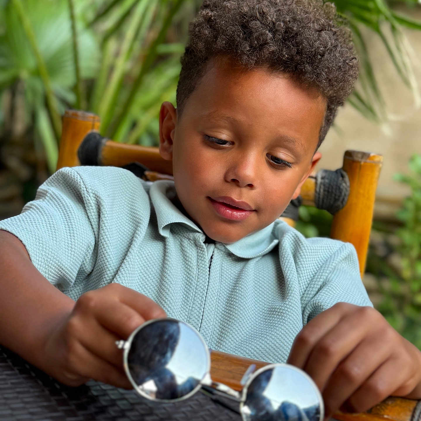 Soann a rejoint le concours — aidez-le/la à gagner de superbes lots ! boy, casual_clothing, child, close_up, curiosity, curly_hair, face, focus, green_plants, hands, leisure, natural_light, outdoor, portrait, reflection, seated, sunglasses, table, wooden_chair, young