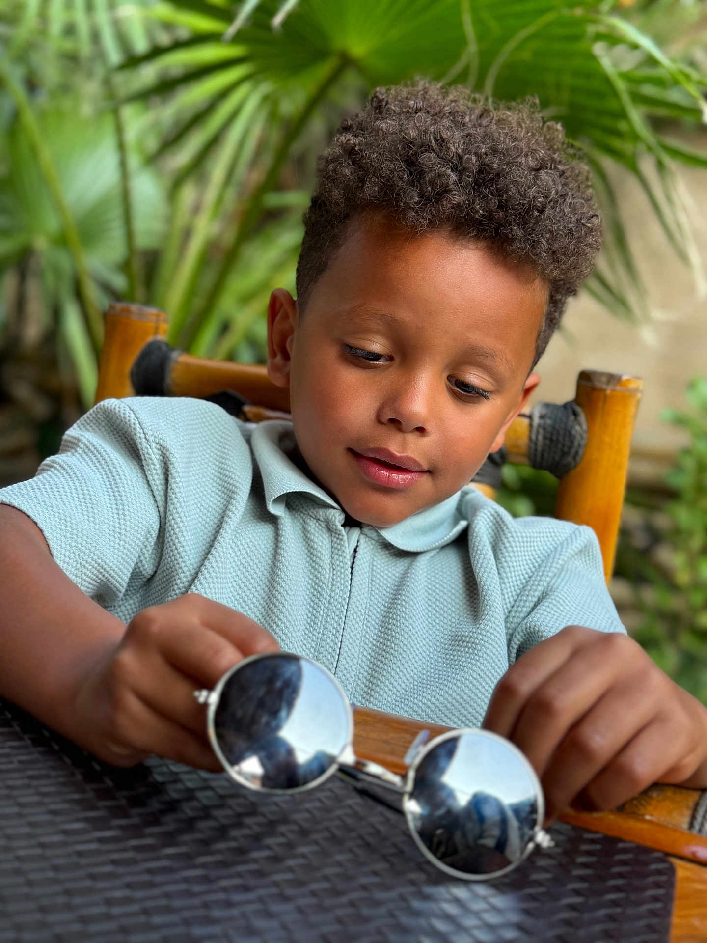 Soann a rejoint le concours — aidez-le/la à gagner de superbes lots ! boy, casual_clothing, child, close_up, curiosity, curly_hair, face, focus, green_plants, hands, leisure, natural_light, outdoor, portrait, reflection, seated, sunglasses, table, wooden_chair, young