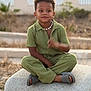 background_blur, boy, casual, child, cross_legged, curly_hair, daylight, fashion, footwear, green_clothing, nature, outdoor, person, portrait, relaxed, shell_necklace, sitting, slippers, stone, young