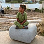 Soann participe au concours pour gagner de l'argent avec cette photo : background_blur, calm, casual_clothing, child, daytime, fence, footwear, grass, green_outfit, nature, outdoor, person, relaxed, residential_area, short_hair, side_view, sitting, stone_block, stroller, urban