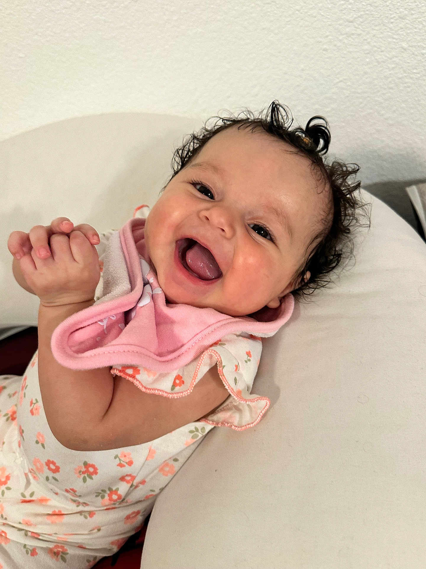 Zeina-Pierina joined the competition — help win amazing prizes! baby, smiling, laughing, curly_hair, pink_bib, floral_clothing, infant, cute, happy, indoors, cushion, white_background, person, child, portrait, closeup, skin, hand, face, lying_down