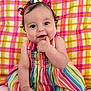 baby, child, smiling, colorful_dress, striped_dress, curly_hair, hair_bows, sitting, plaid_background, indoor, cute, happy, infant, portrait, person, skin, face, hand, foot, bright_colors