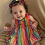 baby, child, smiling, rainbow_striped_dress, colorful_clothing, curly_hair, hair_bows, sitting, beige_blanket, happy, portrait, cute, infant, indoors, playful, young_child, person, bright_colors, closeup, expression