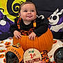baby, background, black_clothing, blanket, child, costume, cute, decoration, face, festive, foot, halloween, hat, holiday, indoor, orange_clothing, pumpkin, sign, smiling, tongue_out