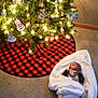 baby, blanket, carpet, christmas_tree, christmas_lights, ornaments, holiday, festive, red_and_black_checkered, tree_skirt, indoor, decorations, infant, floor, cozy, soft_lighting, wrapped, cute, celebration, seasonal