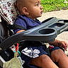 baby, child, stroller, outdoor, sitting, curious, profile, face, eyes, short_hair, blue_shirt, shorts, toy, blanket, snack_tray, grass, sidewalk, sunlight, portrait, infant