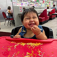 Valentina is registered to the contest to win money with this photo: baby, child, toddler, restaurant, high_chair, smiling, laughing, eating, noodles, food, messy, bib, denim, portrait, indoor, dining_area, table, red, family, happy