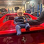 baby, child, car, ferrari, red, garage, luxury_car, automobile, person, man, indoor, lighting, clothing, logo, vehicle, adult, supporting_arm, star_pattern, infant, transportation