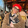 Adèle a rejoint le concours — aidez-le/la à gagner de superbes lots ! woman, dog, french_bulldog, puppy, red_beret, glasses, brown_coat, outdoor_cafe, fairy_lights, flowers, table, chairs, people, warm_lighting, portrait, cute, cozy, winter_clothing, pet, smile