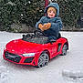 child, snow, toy_car, red_car, winter_clothing, mittens, beanie, hooded_jacket, outdoor, snowfall, greenery, happy, playing, vehicle, small_car, cold_weather, parked_car, snow_covered, fun, automobile