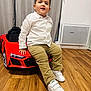 toddler, child, white_shirt, beige_pants, white_shoes, toy_car, red_car, indoor, wooden_floor, curtains, smiling, casual_pose, happy, floor, childrens_toy, person, young_child, playtime, home, cute