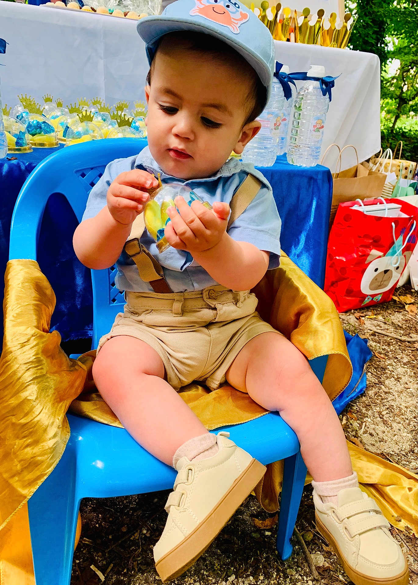 Luis Felipe participe au concours pour gagner de l'argent avec cette photo : toddler, child, blue_cap, blue_shirt, shorts, suspenders, plastic_chair, shoes, outdoor, party_decorations, gift_bags, table, water_bottles, curious, toy, nature, ground, celebration, face, person