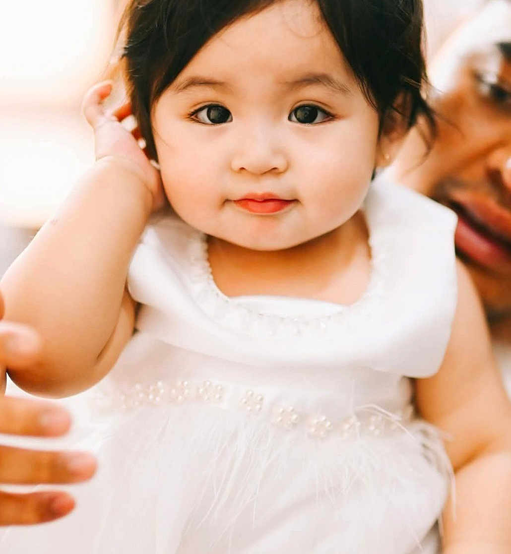 Sofia is registered to the contest to win money with this photo: baby, child, person, white_dress, pearl_details, close_up, portrait, cute, soft_lighting, indoor, hand, adult, supporting_hand, face, smile, curious, black_hair, skin, expression, adorable