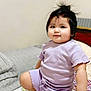 toddler, child, baby, face, smile, hair, purple_clothing, bed, bedspread, floral_pattern, pillow, indoor, skin, hand, foot, cute, person, young_child, clothing, kneeling