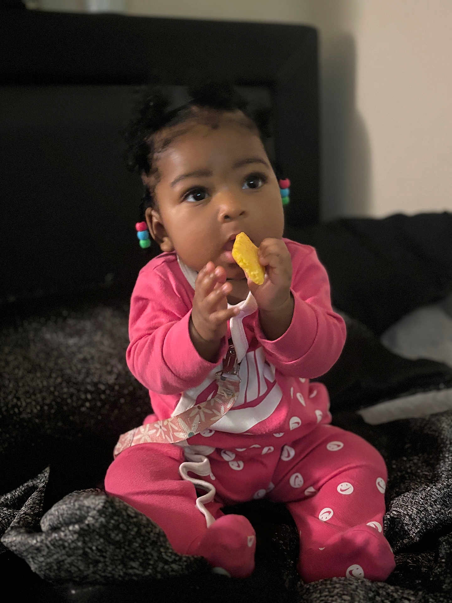 Kyzah joined the competition — help win amazing prizes! baby, child, pink_clothing, smiling_faces_pattern, sitting, bed, blanket, snack, hair_beads, curious_expression, indoors, portrait, cute, young_child, hand_to_mouth, cozy, infant, person, face, clothing