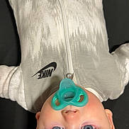 Sunnie is registered to the contest to win money with this photo: baby, infant, pacifier, blue_eyes, onesie, nike, clothing, sleepwear, face, head, cute, child, babywear, lying_down, indoor, soft_lighting, skin, human, portrait, newborn