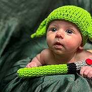 Sunnie is registered to the contest to win money with this photo: baby, infant, knitted_hat, green_hat, toy, knitted_toy, lightsaber, cute, child, portrait, soft_background, fabric, hand, blue_eyes, curious, young_child, indoor, closeup, face, expression
