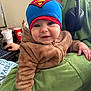 baby, child, superman_hat, brown_jacket, green_hoodie, headphones, indoor, computer_desk, cup, person, smiling, happy, fuzzy_clothing, hand, face, blue_hat, window_light, cozy, casual, young_child