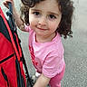 child, toddler, curly_hair, pink_clothing, stroller, outdoor, pavement, smile, person, young_child, casual, holding, walking, cute, daylight, shoes, playful, happy, portrait, expression