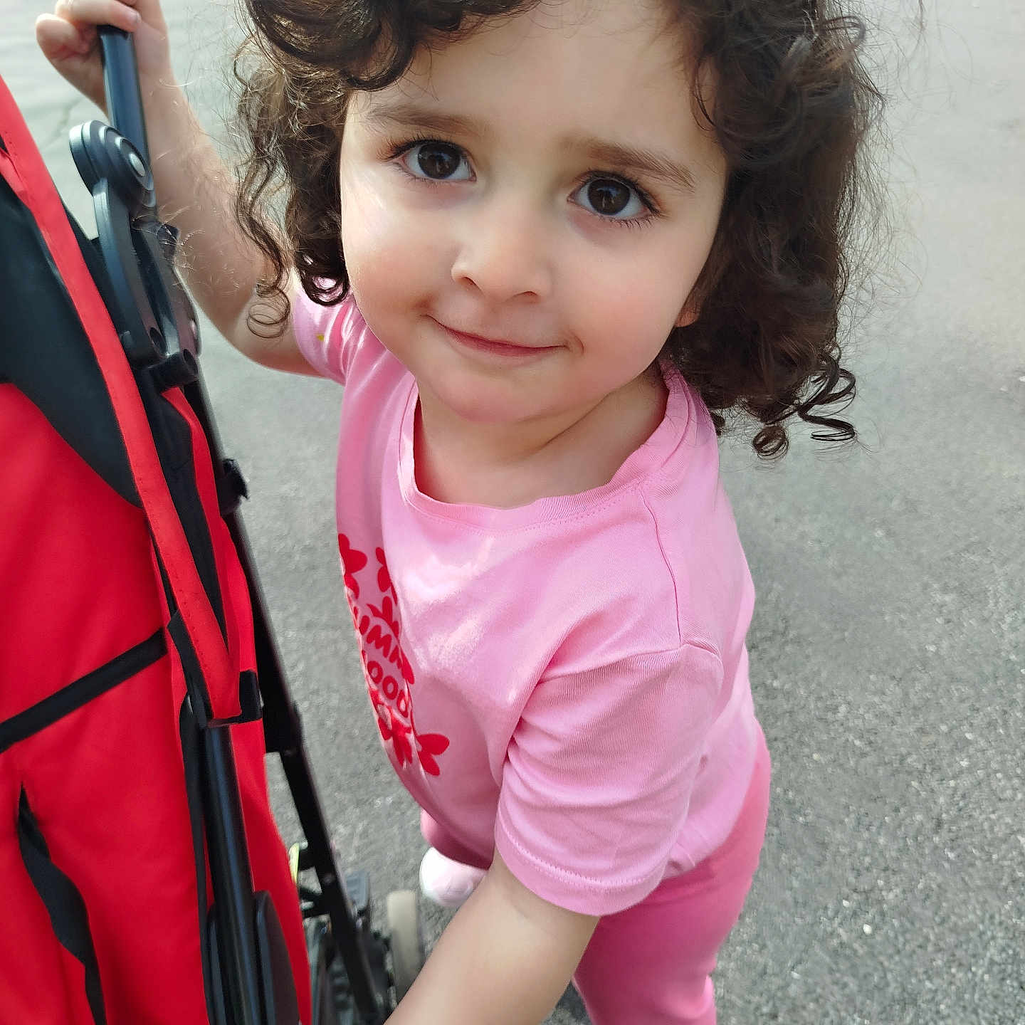 Lina a rejoint le concours — aidez-le/la à gagner de superbes lots ! child, toddler, curly_hair, pink_clothing, stroller, outdoor, pavement, smile, person, young_child, casual, holding, walking, cute, daylight, shoes, playful, happy, portrait, expression