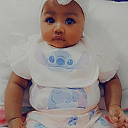 Tessa is registered to the contest to win money with this photo: baby, infant, child, headband, bow, clothing, onesie, bib, stitch_character, bed, blanket, soft_toys, cute, portrait, sitting, indoors, skin, face, hands, expression