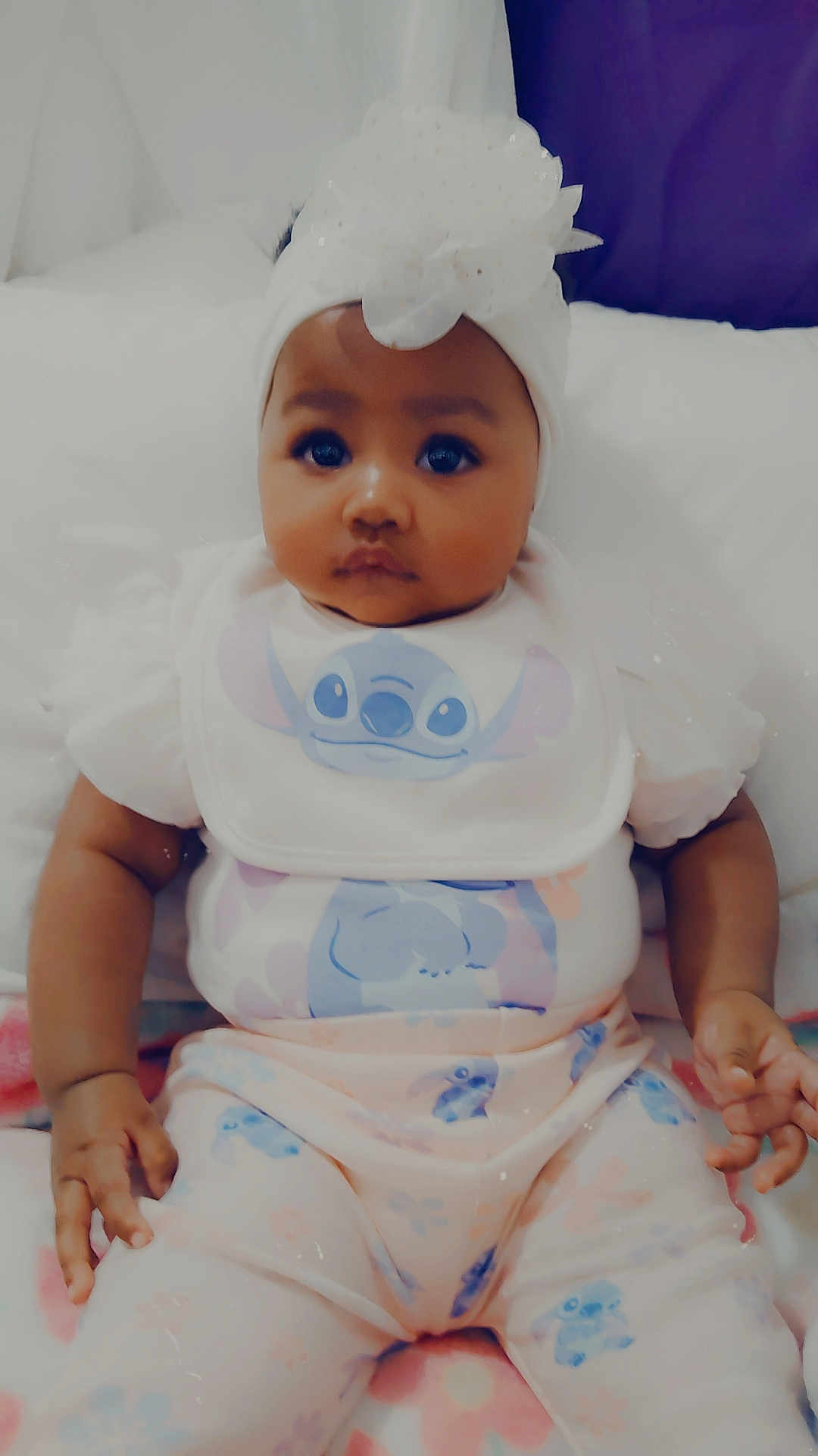 Tessa is registered to the contest to win money with this photo: baby, infant, child, headband, bow, clothing, onesie, bib, stitch_character, bed, blanket, soft_toys, cute, portrait, sitting, indoors, skin, face, hands, expression