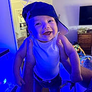 Roman is registered to the contest to win money with this photo: baby, child, infant, smiling, happy, portrait, cap, hat, being_held, adult_hands, indoor, living_room, television, dresser, toy, blue_lighting, clothing, playful, close_up, photography