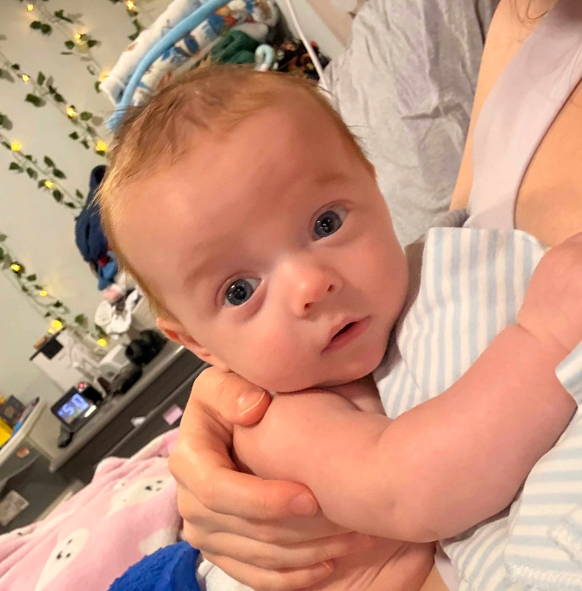 Harlan is registered to the contest to win money with this photo: baby, infant, child, person, holding, arm, striped_clothing, face, eyes, hand, indoor, blanket, bed, adult_hand, furniture, fairy_lights, curious_expression, skin, closeup, portrait