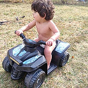 Alexander is registered to the contest to win money with this photo: child, toddler, outdoor, backyard, toy_vehicle, quad_bike, ride_on_toy, curly_hair, profile, sitting, grass, autumn, leaves, fence, playtime, recreation, vehicle, handlebars, bare_chested, daylight