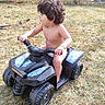 child, toddler, outdoor, backyard, toy_vehicle, quad_bike, ride_on_toy, curly_hair, profile, sitting, grass, autumn, leaves, fence, playtime, recreation, vehicle, handlebars, bare_chested, daylight