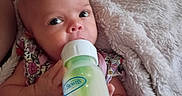 Ila is registered to the contest to win money with this photo: baby, infant, child, bottle, feeding, blanket, floral_outfit, blue_eyes, wide_eyed, lying_down, indoors, portrait, close_up, adult_hand, soft_blanket, cozy, baby_care, milk_bottle, family, safe