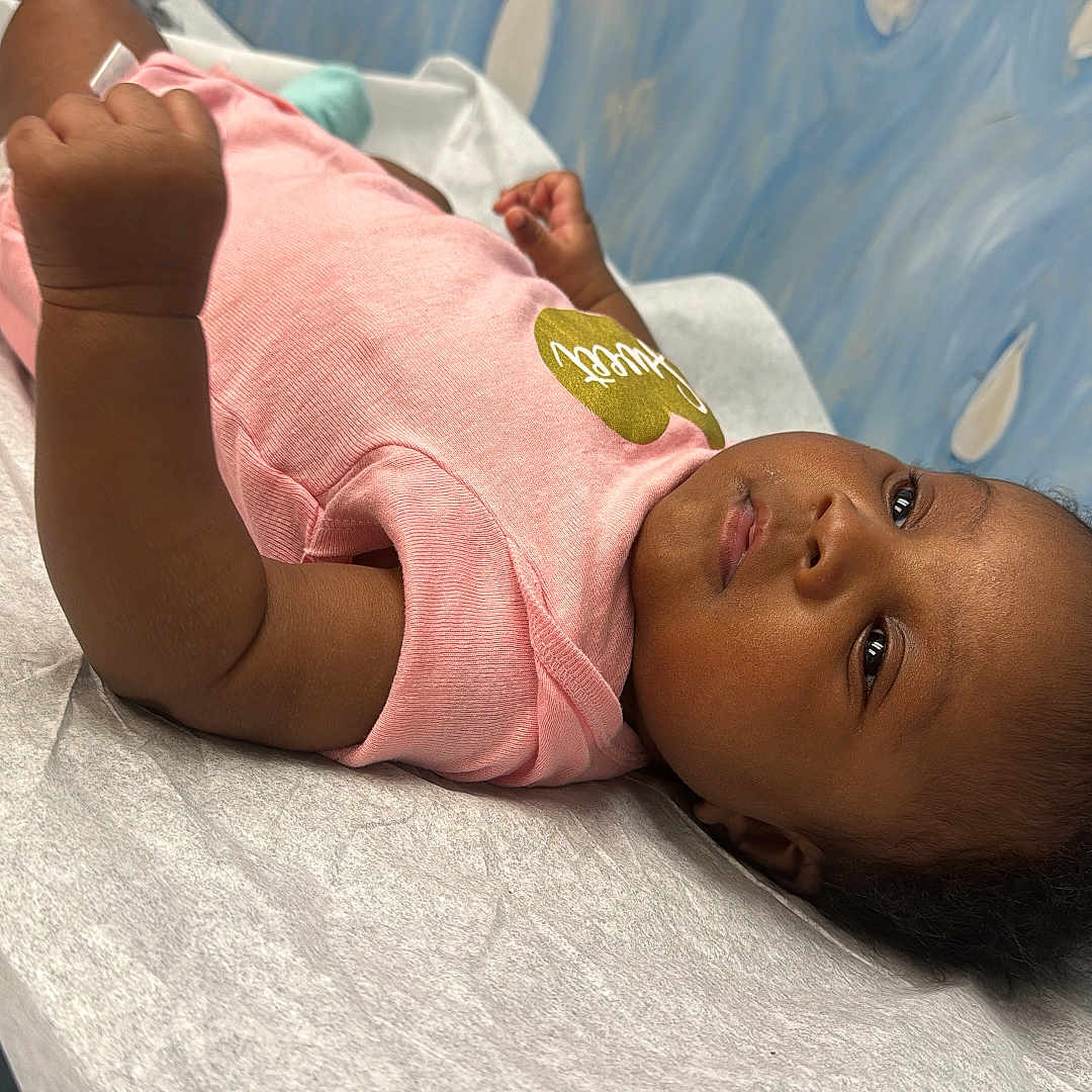 Tochi is registered to the contest to win money with this photo: arm, baby, blue_wall, casual_clothing, child, hair, hand, healthcare, indoors, infant, looking_sideways, lying_down, medical_table, paper_cover, patient, pink_shirt, portrait, skin, teardrop_pattern, young_child