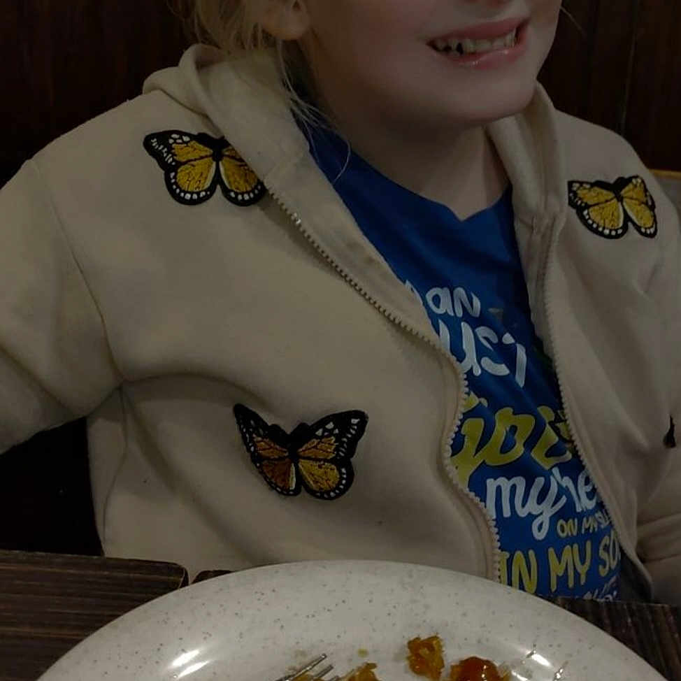 Angela is registered to the contest to win money with this photo: appetite, blonde_hair, butterfly_patch, casual_clothing, child, dining, face, food, fork, happy, indoor, jacket, meal, person, plate, restaurant, seated, smiling, table, young