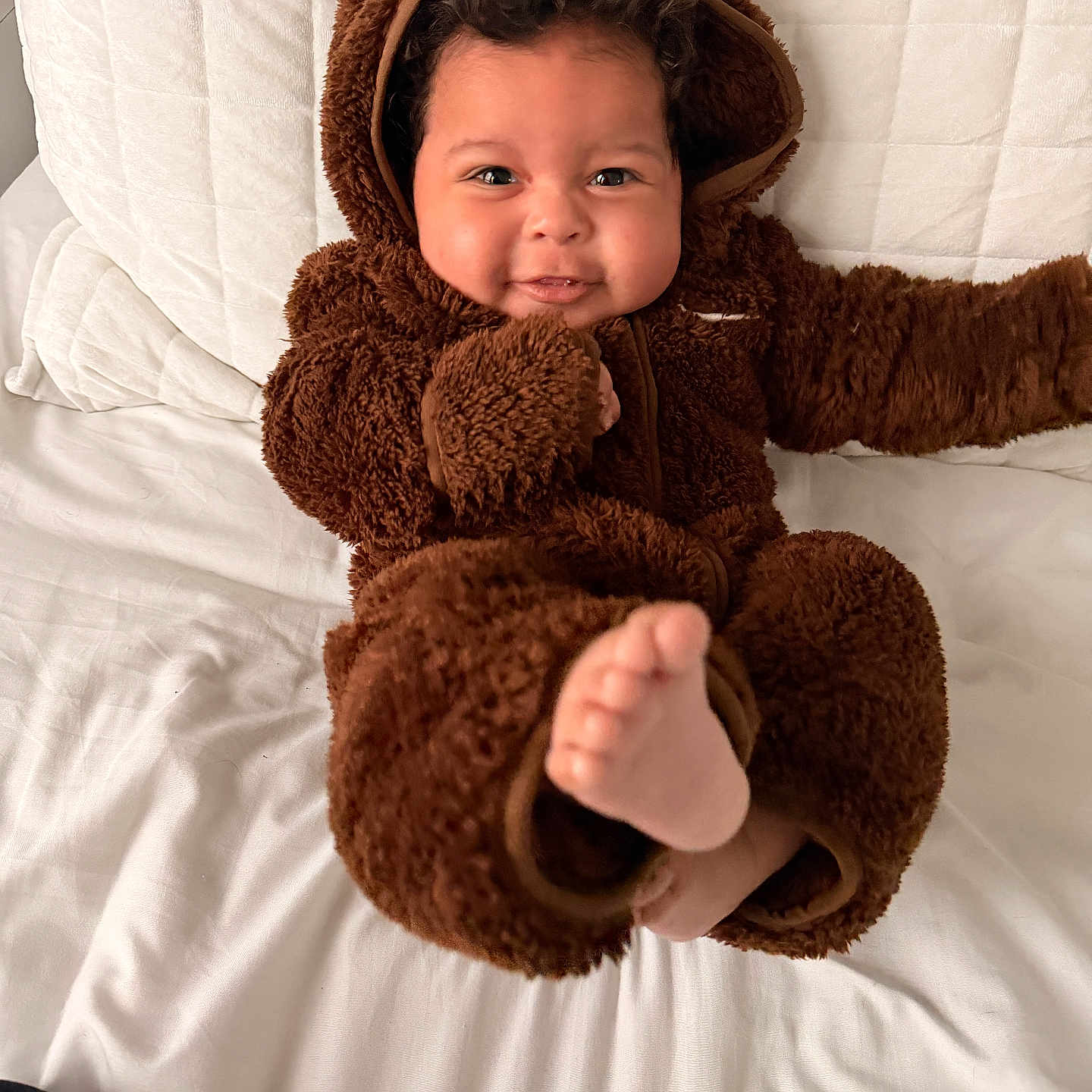 Raian is registered to the contest to win money with this photo: baby, child, fuzzy_clothing, brown_onesie, cute, smiling, foot, bed, white_bedspread, indoor, portrait, childhood, happy, playful, infant, cozy, warm_clothing, face, person, soft_texture