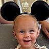 adult, baby, beaded_necklace, blonde_hair, blue_eyes, bows, child, closeup, earrings, face, happy, headband, indoor, mickey_mouse_ears, portrait, sitting, smiling, toddler, vehicle, white_outfit