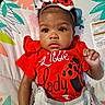 Phor'Ever is registered to the contest to win money with this photo: baby, infant, child, portrait, lying_down, bow, headband, red_outfit, ladybug, patterned_clothing, smiling, wide_eyes, curly_hair, dark_skin, hand, blanket, seat, colorful_background, indoor, photography