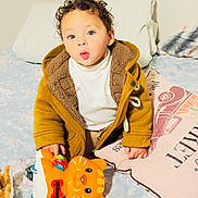 Isaac is registered to the contest to win money with this photo: toddler, child, toy, orange, cat_toy, jacket, brown_jacket, curly_hair, bed, pillow, plush_toy, indoor, person, clothing, white_shirt, cute, expression, furniture, home, young_child