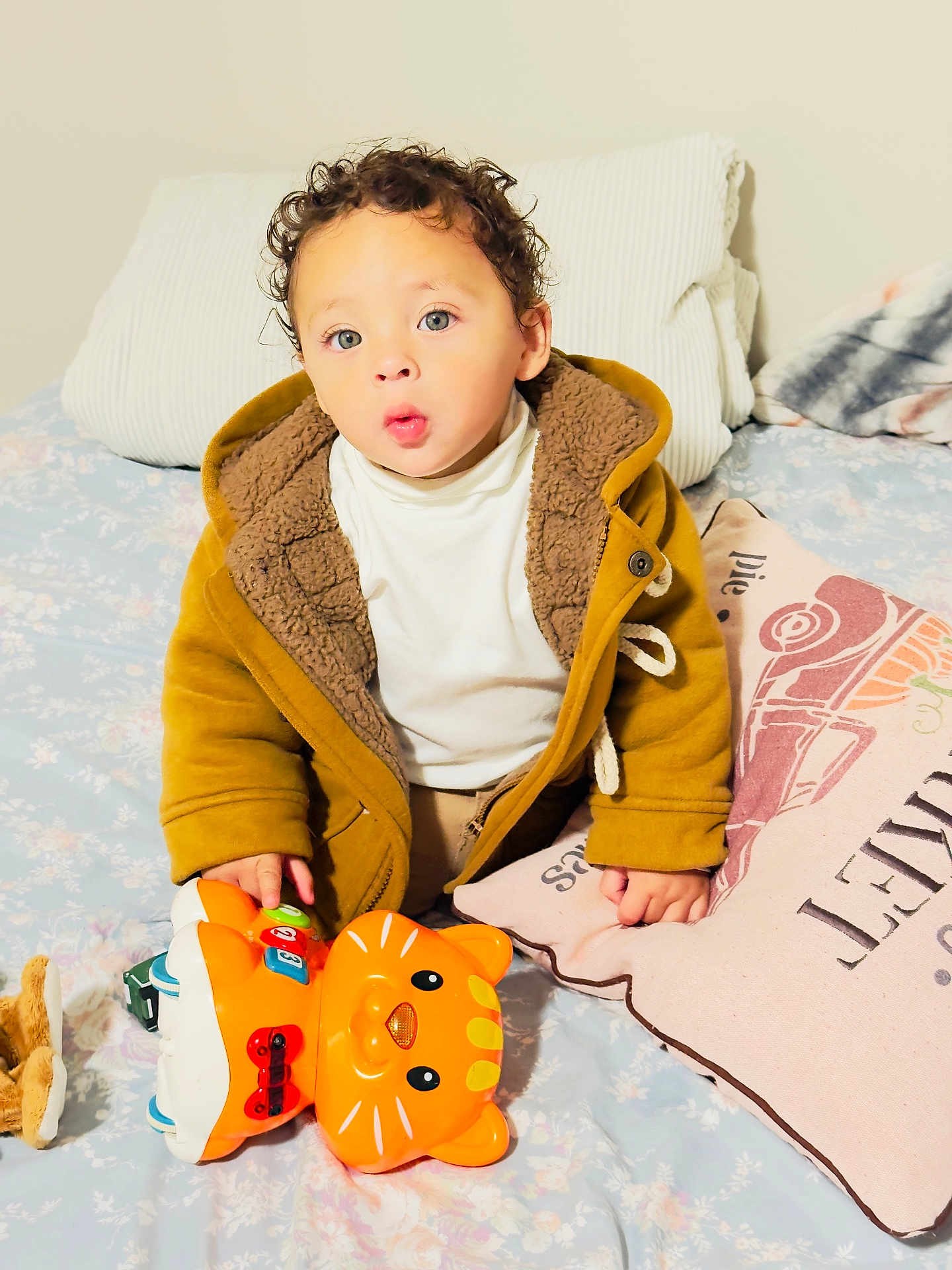 Isaac is registered to the contest to win money with this photo: toddler, child, toy, orange, cat_toy, jacket, brown_jacket, curly_hair, bed, pillow, plush_toy, indoor, person, clothing, white_shirt, cute, expression, furniture, home, young_child