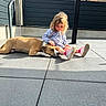 child, girl, dog, pet, outdoor, patio, sunlight, sitting, lying_down, petting, friendship, companion, playful, blonde_hair, dress, sneakers, residential, fence, paving_stones, portrait