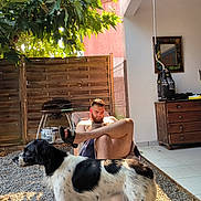 Sam participe au concours pour gagner de l'argent avec cette photo : man, dog, outdoor, sunlight, fence, tree, grill, chair, phone, beard, relaxing, tile_floor, wooden_dresser, shade, leafy_tree, black_and_white_dog, casual, summer, backyard, sandals