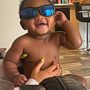 Symere is registered to the contest to win money with this photo: baby, child, sunglasses, smiling, happy, hand, nails, indoor, kitchen, person, skin, face, furniture, floor, reflection, holding, casual, cute, infant, human