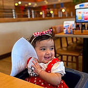 Lyna Marwah is registered to the contest to win money with this photo: baby, child, smiling, high_chair, red_dress, polka_dot, headband, paper, restaurant, indoor, table, wooden_chair, happy, cute, person, toddler, seated, face, clothing, portrait