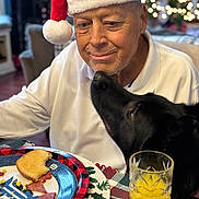 Rex is registered to the contest to win money with this photo: elderly_man, santa_hat, black_dog, breakfast, toast, orange_juice, table, plate, holiday, christmas_lights, indoor, affection, smile, plaid, napkin, white_shirt, cozy, face, pet, warm_lighting