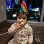 toddler, child, party_hat, birthday, cake, frosting, messy, indoors, floor, night, city_lights, blurred_background, cute, sitting, clothing, celebration, small_child, face, hand, fun