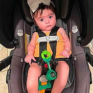 Amara is registered to the contest to win money with this photo: baby, infant, child, car_seat, bow, headband, orange_clothing, harness, toy, green_toy, sitting, portrait, wide_eyes, expression, indoor, vehicle, safety, baby_carrier, bare_feet, photography