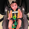 baby, infant, child, car_seat, bow, headband, orange_clothing, harness, toy, green_toy, sitting, portrait, wide_eyes, expression, indoor, vehicle, safety, baby_carrier, bare_feet, photography