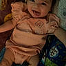baby, bedding, blanket, casual_photo, child, dark_lighting, eyes, face, happy, indoor, infant, lying_down, mouth, patterned_fabric, peach_outfit, pillow, portrait, smartphone_screenshot, smiling, social_media
