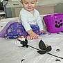 baby, child, purple_skirt, headband, ears, toy_spiders, halloween_bucket, bedspread, room, curious, sitting, foot, white_clothing, indoor, playful, decorations, toys, smiling, cute, costume